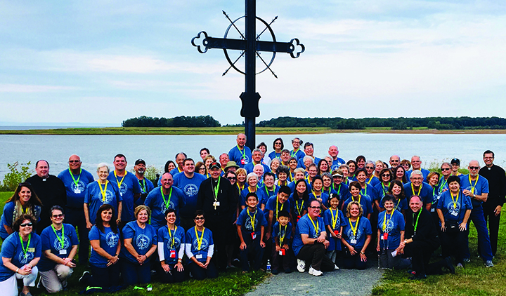A large group of pilgrimes poses in front of a tall metal cross, known as the Deportation Cross in Nova Scotia. 